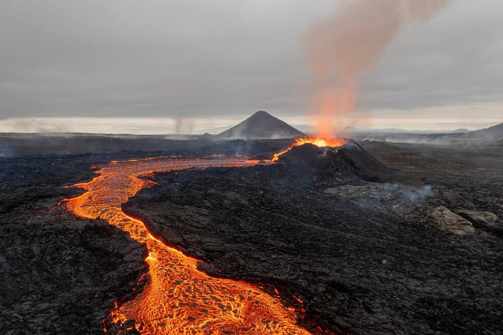 La lave chaude s'écoulant à travers une étendue de roches volcaniques sombres, avec un volcan fumant à l'arrière-plan.