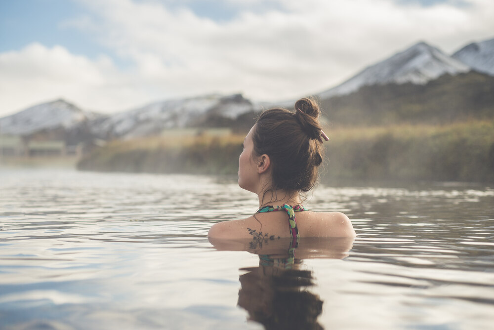 Une femme profitant des eaux d'une source chaude naturelle en Islande, avec un paysage montagneux en arrière-plan.