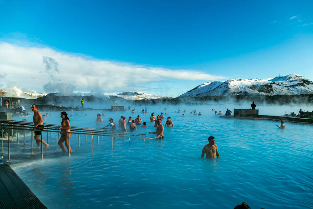 Des baigneurs profitant de la célèbre lagune bleue, entourés de vapeur et de montagnes enneigées sous un ciel bleu.