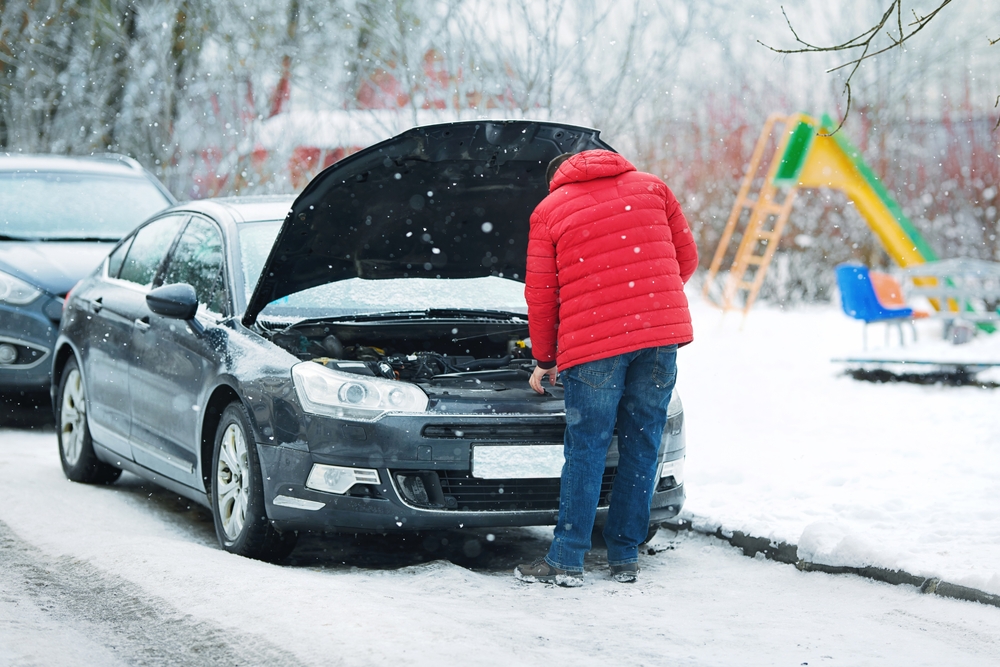 Un homme en manteau rouge inspecte le moteur d’une voiture en panne sous la neige, capot ouvert sur une route glacée près d’un parc