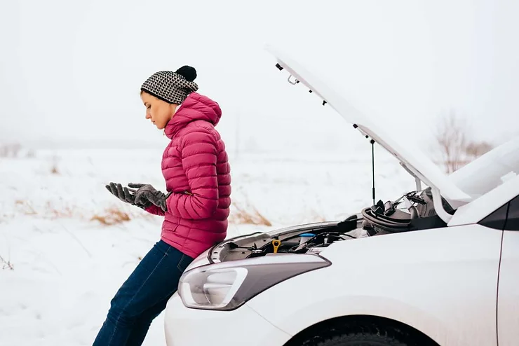 Une femme en manteau rose regarde son téléphone à côté d'une voiture en panne avec le capot ouvert sur une route enneigée