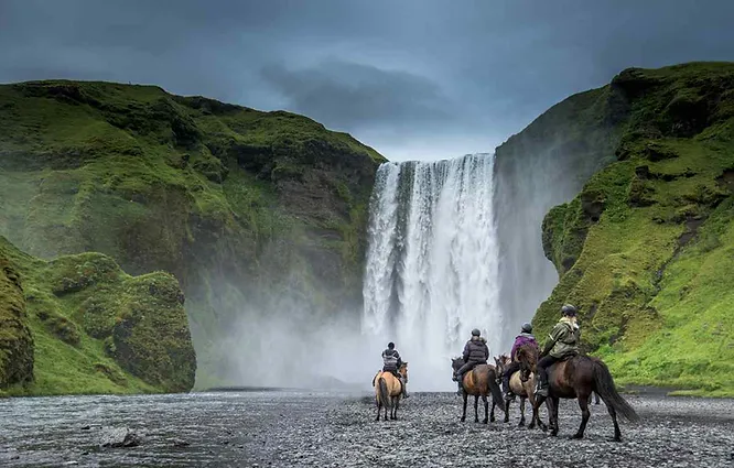 Cavaliers sur des chevaux islandais approchant la cascade de Skógafoss en Islande, entourée de falaises verdoyantes sous un ciel nuageux.