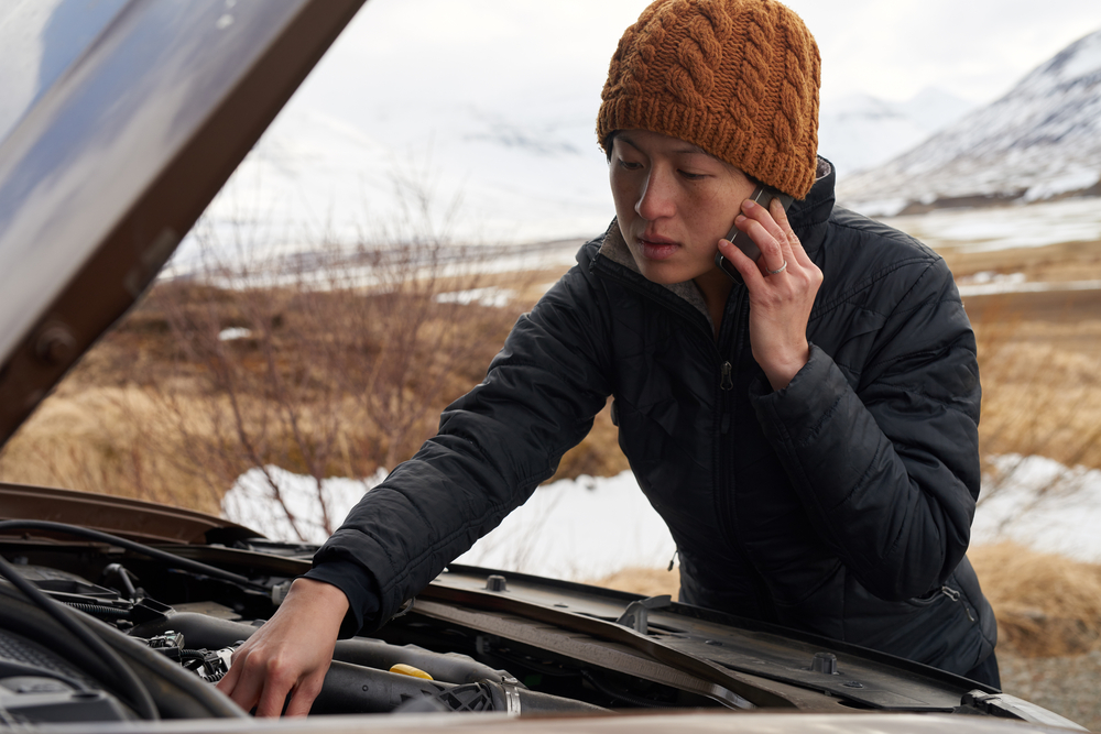 Femme en bonnet orange appelant de l’aide au bord de la route en Islande, capot ouvert face à une panne mécanique.