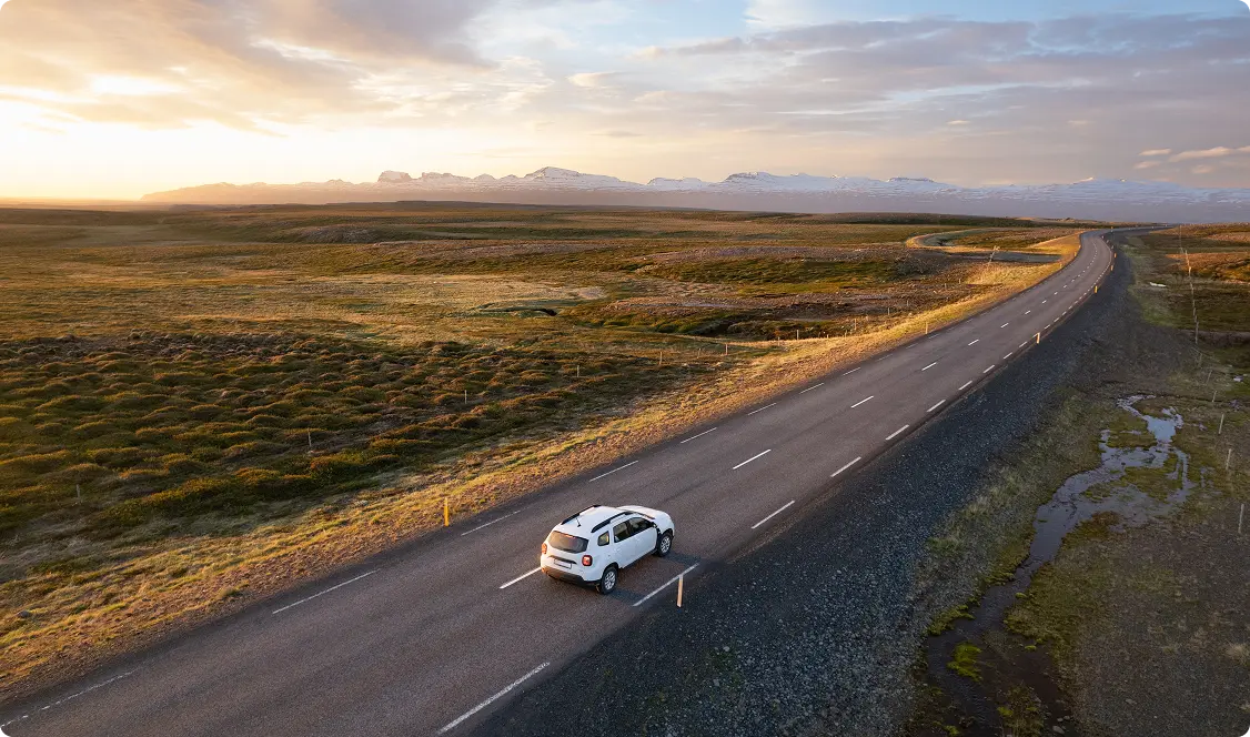 Vue aérienne d'un fleuve sinueux et des hautes terres accidentées de l'Islande, avec une voiture traversant un pont, symbolisant l'aventure et la liberté d'explorer l'Islande avec une voiture de location.